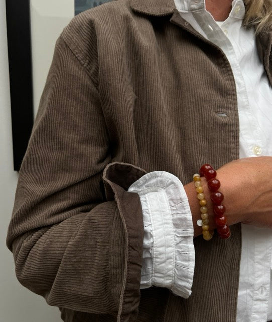 Frau trägt braune Cordjacke und weiße Bluse mit zwei Perlenarmbändern von Soi aus roter und gelber Jade am Handgelenk.